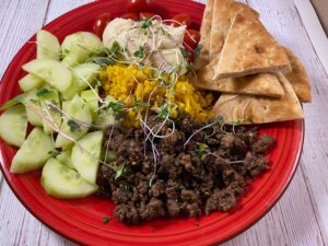 red plate with pita, cucumber, tomato, tzatziki, hummus, turmeric rice, microgreens and ground lamb