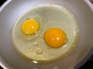 Two eggs cooking in a skillet, showing a dark orange farm fresh yolk next to a pale yellow store-bought yolk.